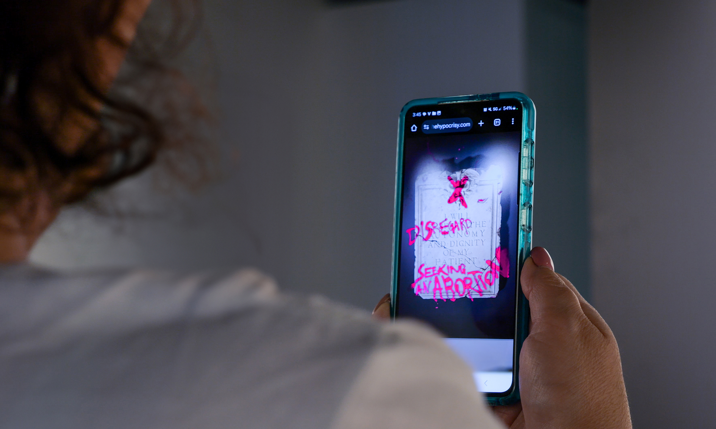 A person holds up their phone to scan an image with Saddington Baynes' AR app for the No More Hippocrites campaign. The image on the screen is a shot of the hippocratic oath with graffiti on top. It reads "I will disregard the autonomy and dignity of my patient seeking an abortion."